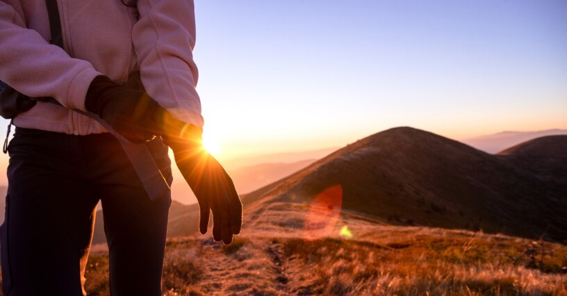 Eine Person zieht Trekkinghandschuhe in einer bergigen Landschaft bei Sonnenaufgang an