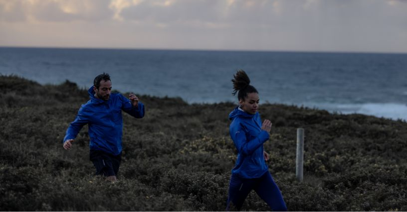 Zwei Personen joggen am Meer in blauen Sportjacken