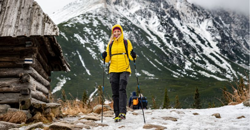 Eine Person wandert im Schnee mit Stöcken vor einer Holzhütte und Bergen im Hintergrund