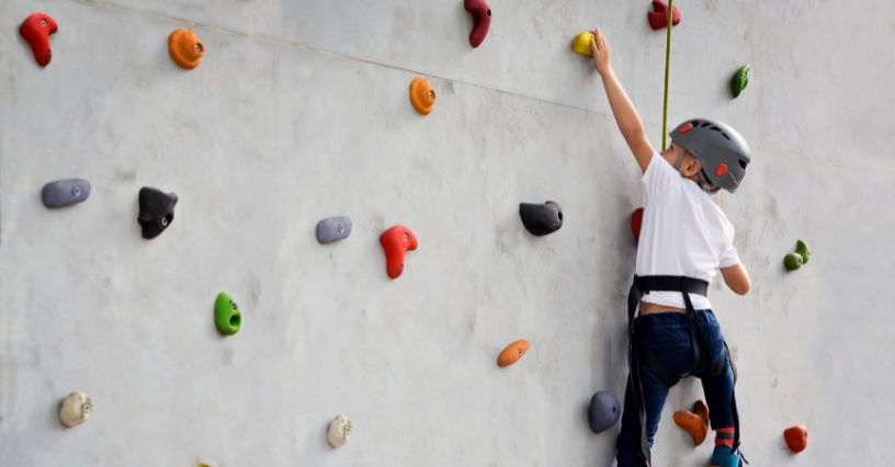 Ein Kind klettert an einer Kletterwand mit bunten Griffen, gesichert durch Klettergurt und Helm