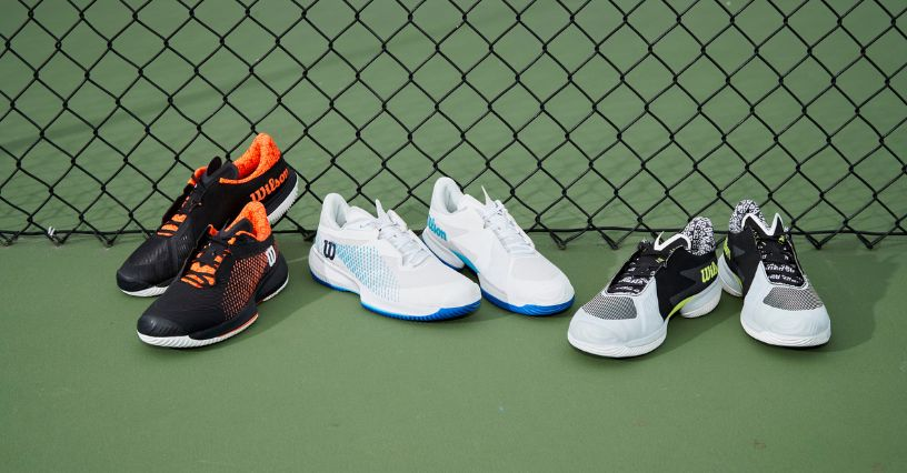 Drei Paar Tennisschuhe stehen vor einem Zaun auf einem Tennisplatz