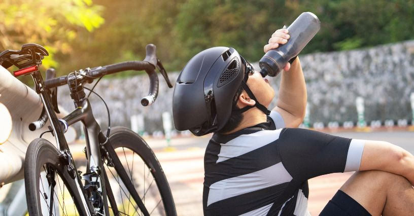 Eine Person trägt einen Fahrradhelm und trinkt aus einer Wasserflasche neben einem Rennrad