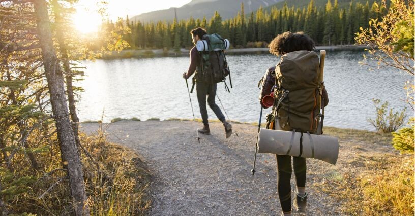 Zwei Personen wandern mit Trekkingrucksäcken an einem See entlang