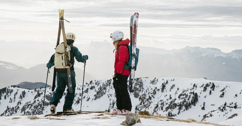 Zwei Personen stehen mit Skiern und Rucksäcken auf einem verschneiten Berggipfel