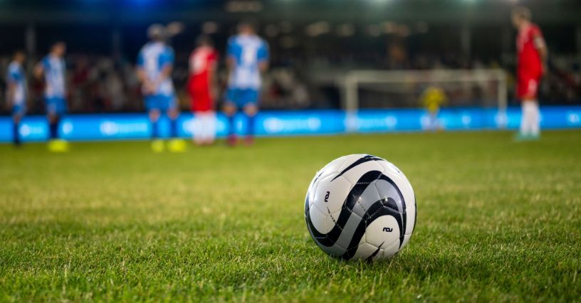 Ein Fußball liegt auf dem Spielfeld, im Hintergrund sind Spieler zu sehen