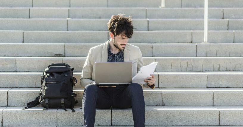 Mann sitzt auf einer Treppe mit Laptop und Dokumenten neben einem Rucksack
