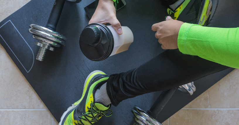 Eine Person hält einen Shaker und sitzt auf einer Trainingsmatte mit Hanteln
